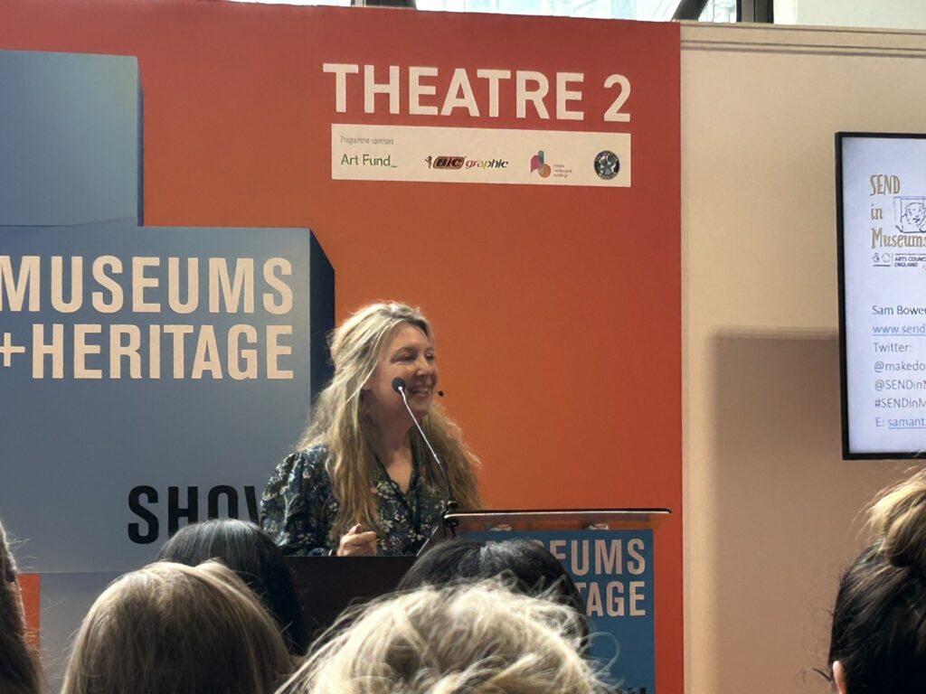 Photo of Sam Bowen a white woman with long blonde hair waring a blue patterned dress and standing at a lecturn as she speaks at the Museum and Heritage Show