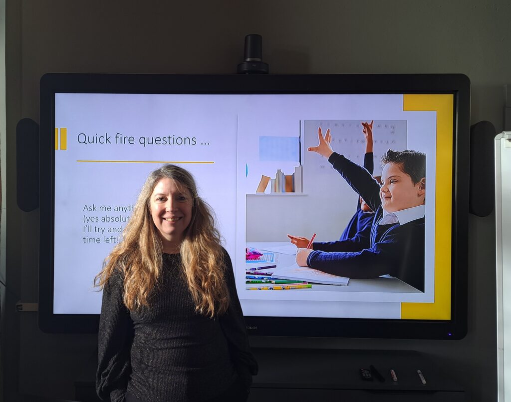 Photo of Sam Bowen a white woman with long blonde hair wearing a black top and smiling. She stands in front of a AV screen with a picture of a boy raising his hand and the words Quick fire questions